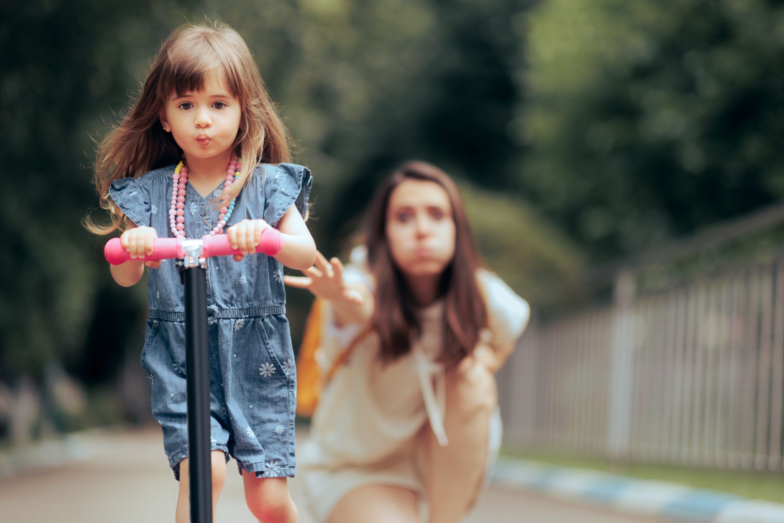 A young girl rides a scooter with focus in a park, donning a denim dress and necklace. In the background, an adult woman reaches out with concern—a subtle nod to the effects of helicopter parents. Blurred greenery and a fence line the path.