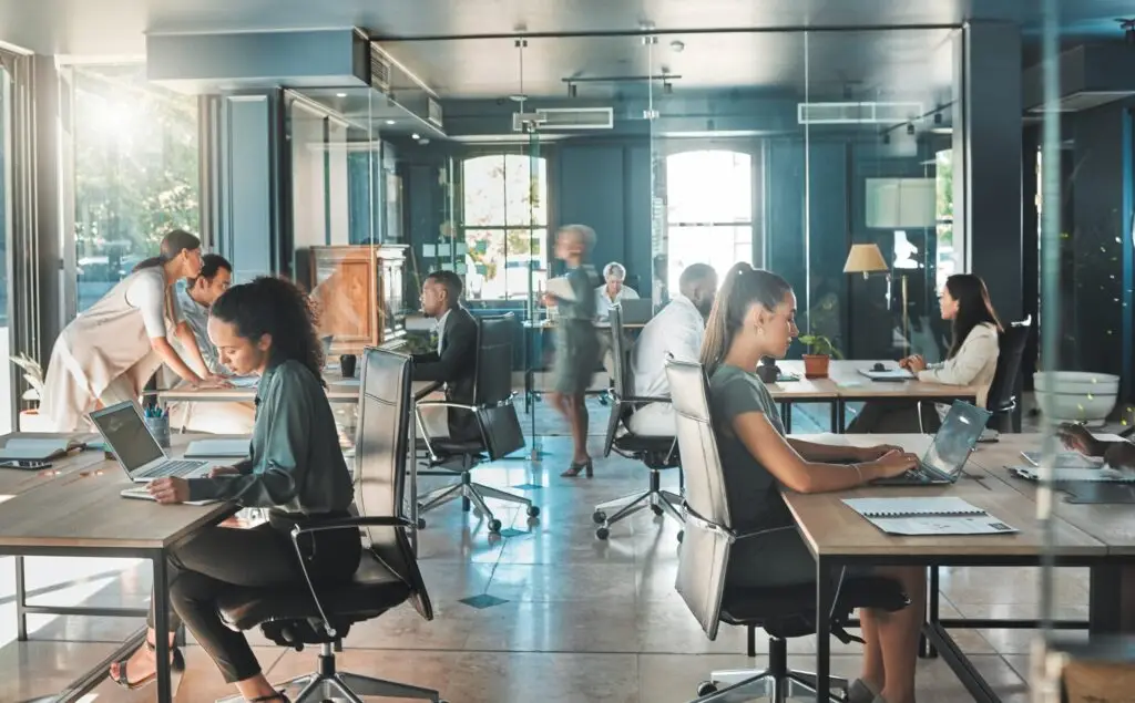 A modern open-plan office buzzes with activity as several people work at desks. Some type on laptops, while others shuffle through papers. Tech seamlessly integrates into their workflow, enhancing productivity. Large windows flood the space with natural light, creating a busy, collaborative atmosphere.