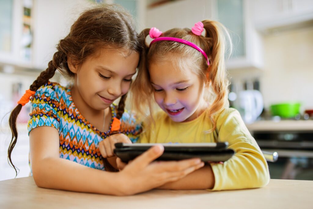 Two young girls, one with braids and the other sporting a pink headband, are smiling and looking at a tablet together in the kitchen. They appear engaged and happy, exploring "Ten of the Best Apps for Child Development" to make learning fun.