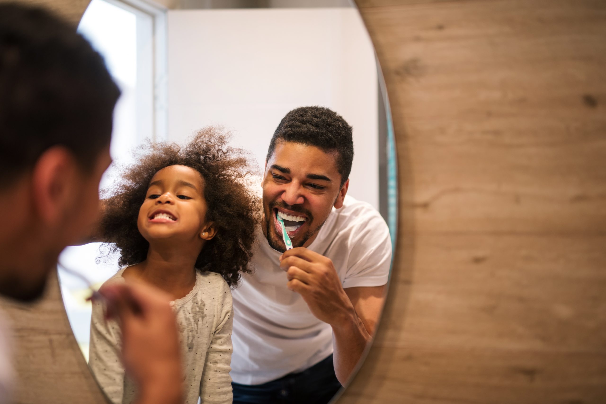 A father and his young daughter smile while brushing their teeth together in front of a round mirror, creating a joyful moment that highlights the benefits of routines for kids. The well-lit bathroom features wooden accents, setting the stage for this playful start to their day.