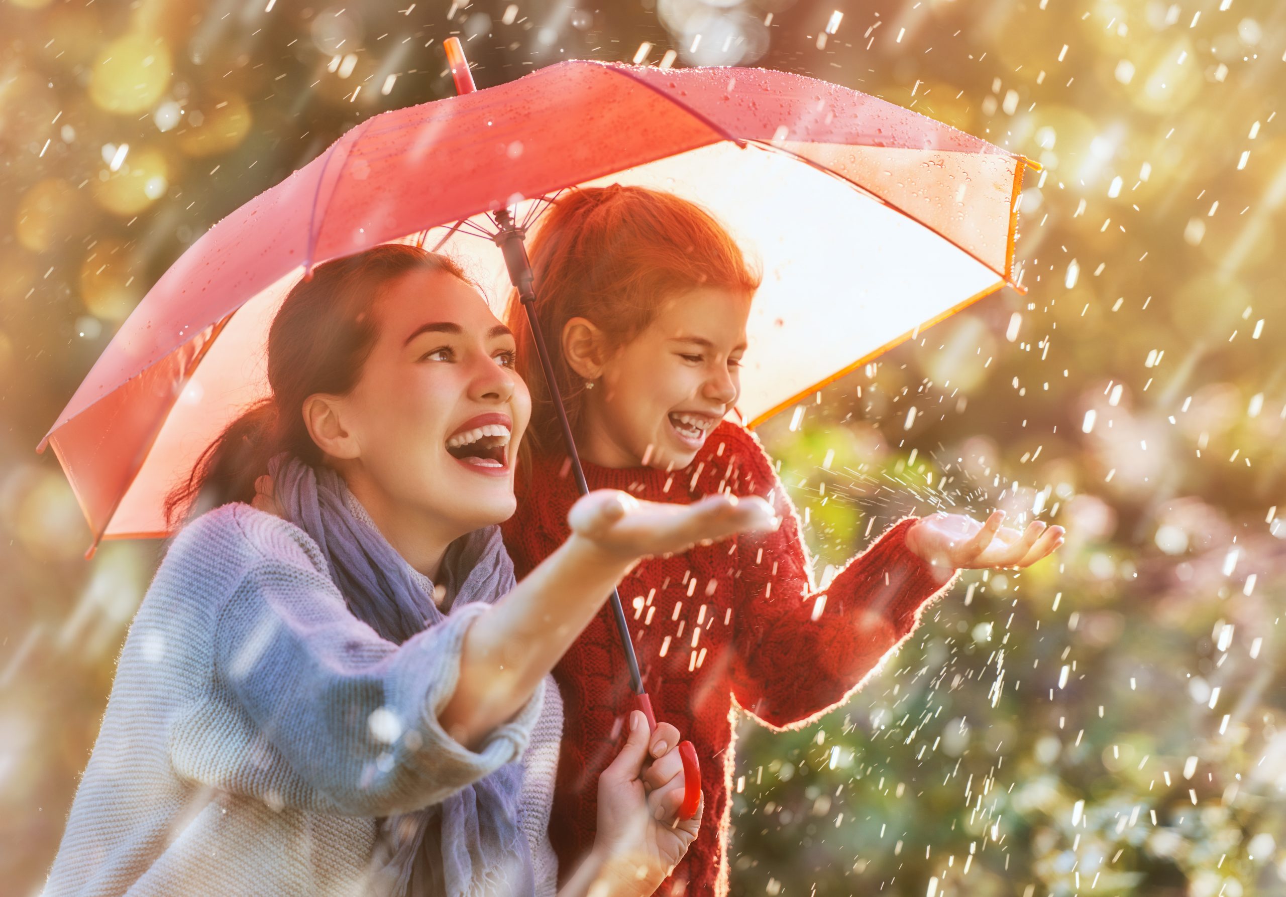 Two people holding a red umbrella, laughing as they explore one of the "One Hundred Rainy Day Activities for Kids," reaching out to catch raindrops. The scene is warm and bright, with sunlight filtering through the trees in the background, capturing the joy of enjoying a rainy day outdoors.