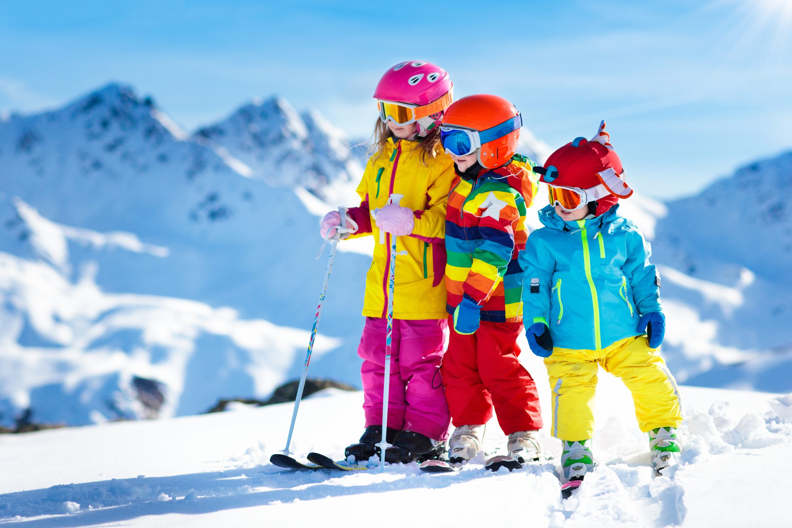 Three children in colorful ski outfits and helmets stand on a snowy mountain slope, pondering "To Ski or Not to Ski: Winter Sports for Kids." They hold ski poles against a backdrop of snowy peaks and a clear blue sky.