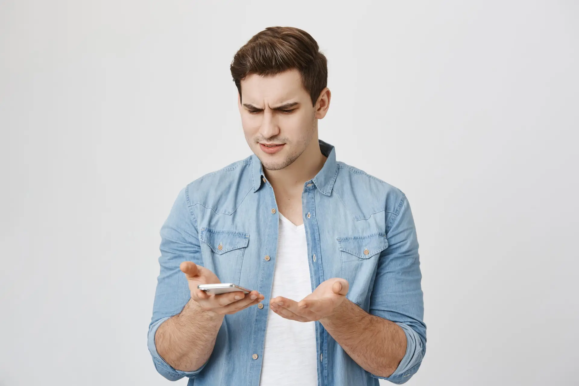 A man in a denim shirt stands against a plain background, looking confused as he examines a smartphone in his hand. His other hand is slightly raised, palm up, suggesting frustration or uncertainty—perhaps he's pondering how to avoid arguments online while fighting over text.