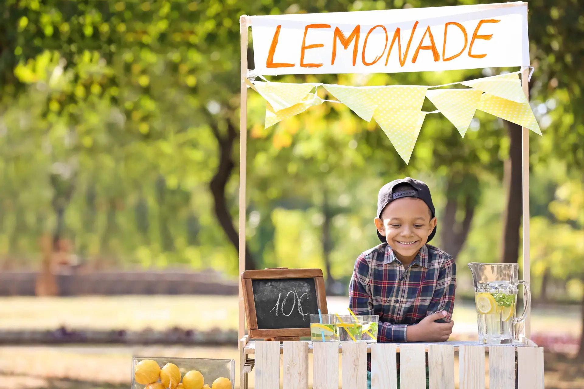 A young boy in a plaid shirt and backward cap smiles at his lemonade stand in the park. The budding entrepreneur showcases one of the best business ideas for kids, with yellow bunting and a chalkboard sign displaying "Lemonade 10¢" next to a glass pitcher on the counter.