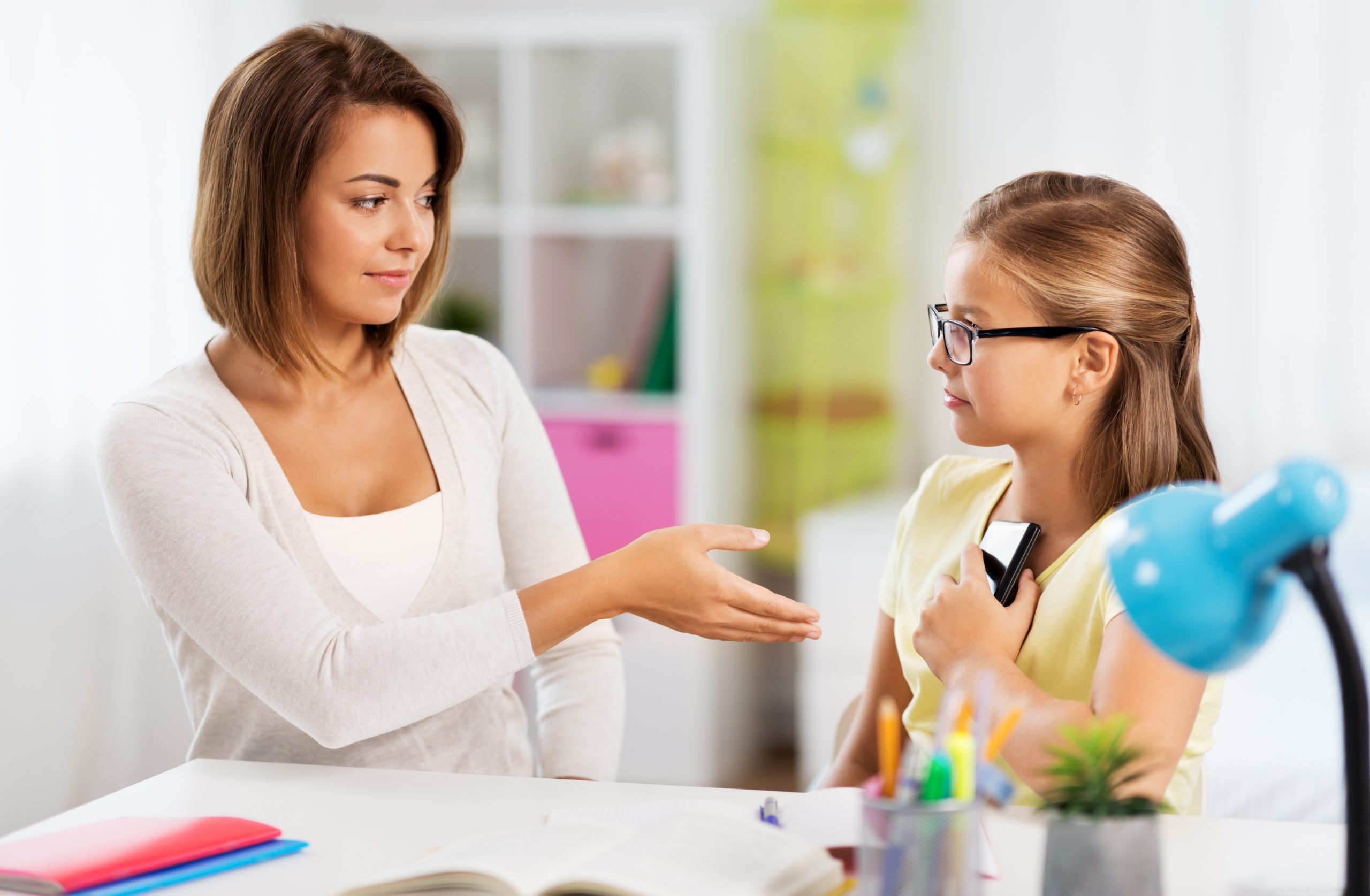 A woman with short brown hair gestures towards a young girl with glasses, who holds a book close to her chest. Seated at a table with stationery, in a brightly lit room, they seem to be discussing whether parents should take away phones as punishment.