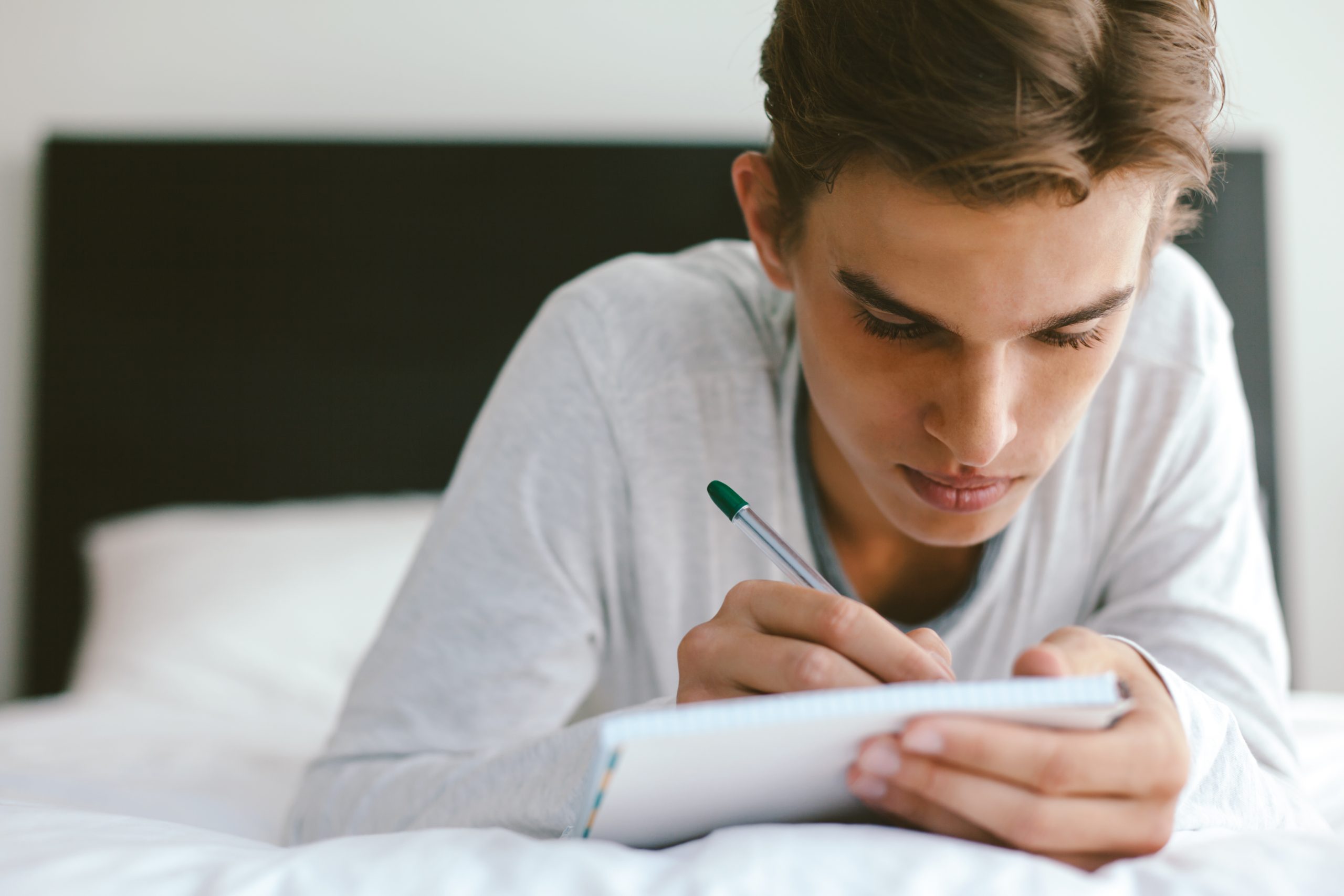 A young person with short hair lies on a bed, focused on journaling their family history in a notebook. Wearing a light-colored long-sleeve shirt, they look serene amid the white sheets and softly lit room—a moment of reflection in an age where using tech often guides such journeys.