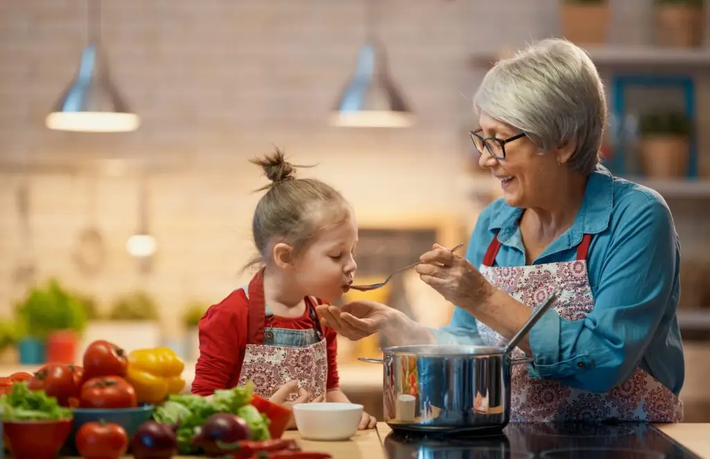 A grandmother and her granddaughter are joyfully engaged in the kitchen. The grandmother, donning glasses and an apron, feeds the little girl a spoonful of food from a pot. Surrounded by fresh vegetables, they relish the joy of cooking together—never burdened by the rigid rules of clean eating.