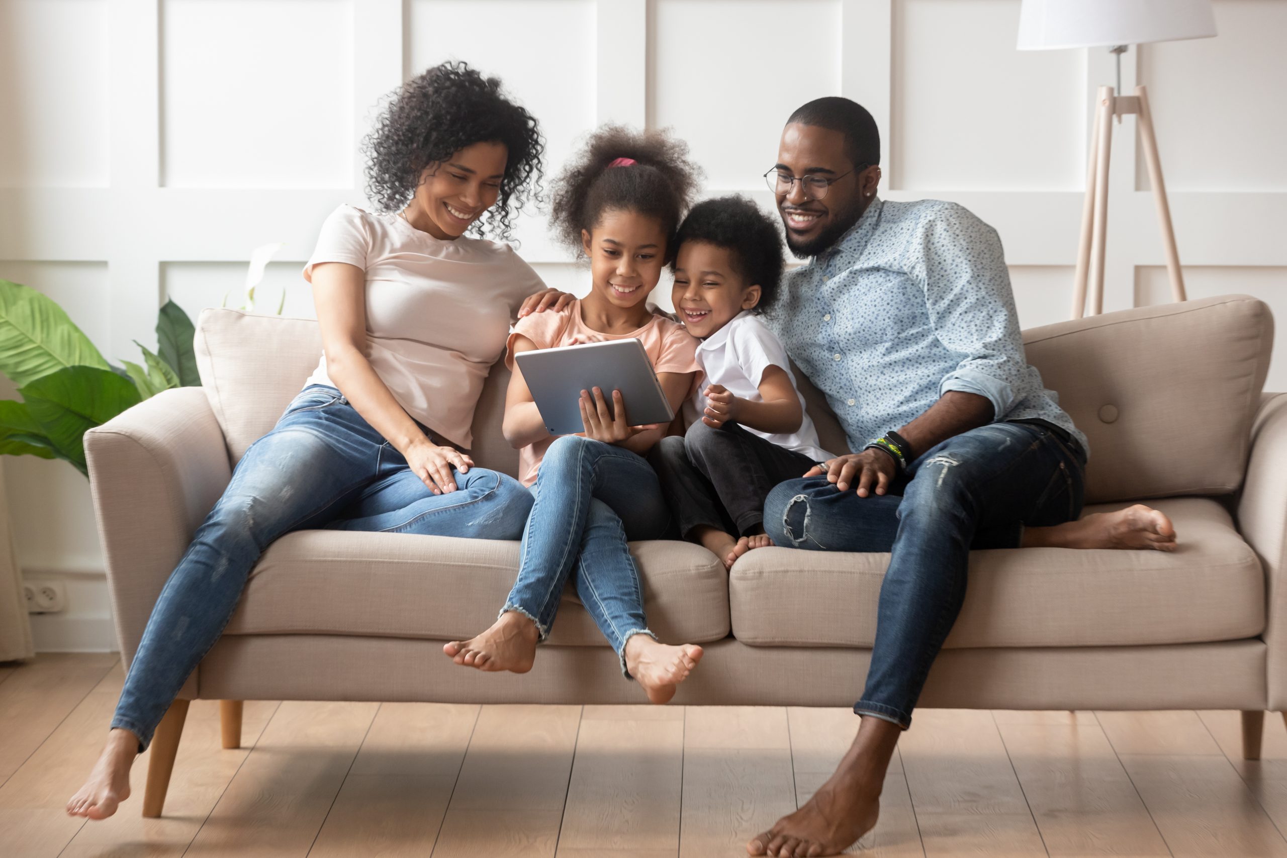 A family of four sits on a sofa, happily engaged with YouTube Kids on a tablet. The parents are on either side of their two children, who are settled in the middle. The room is bright with natural light and a plant visible in the background.