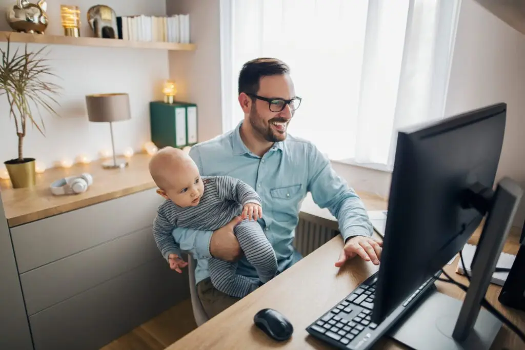 A man working from home on a computer while holding a baby, perhaps researching the top 10 parenting groups online. The well-lit room features a modern interior with shelves, books, and decorative lights. He is smiling, wearing glasses and a blue shirt, balancing work and family life seamlessly.