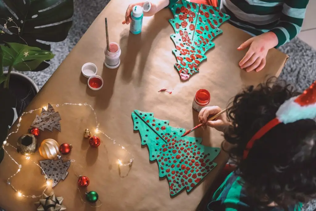 Children painting cardboard Christmas trees at a craft table, surrounded by fairy lights and ornaments, creating a festive atmosphere. One child dons a Santa hat, adding to the holiday spirit and inspiring ideas for 20 easy homemade Christmas gifts.