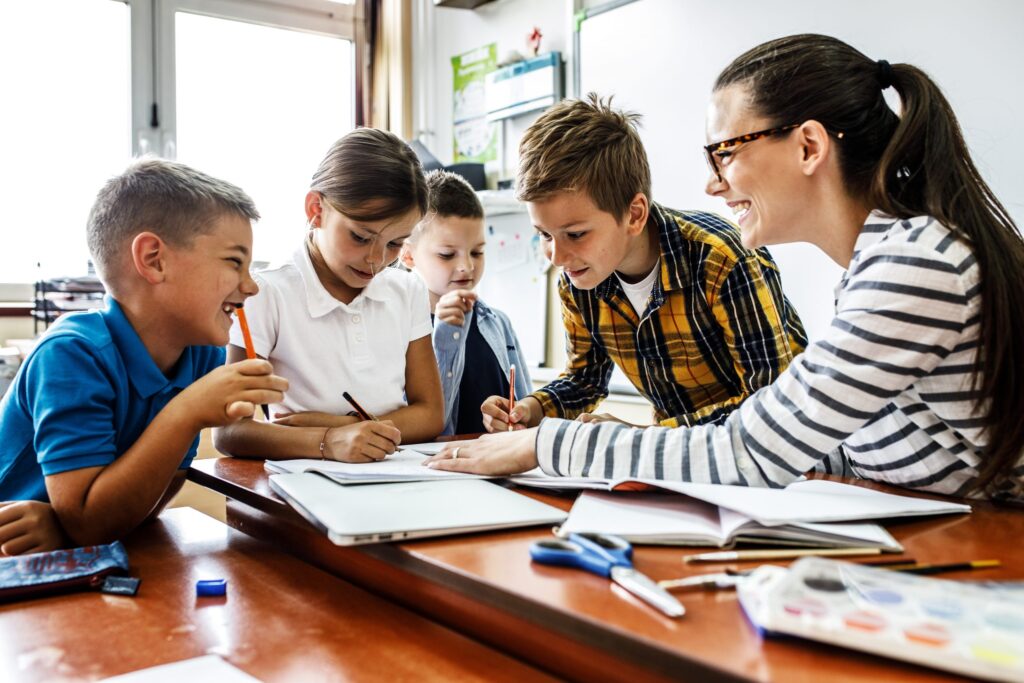 In a tech-free classroom, a teacher sits at a table with four smiling children, engaging in a group activity. The children are focused on their workbooks, exchanging ideas. The bright classroom buzzes with creativity as art supplies are scattered on the table.