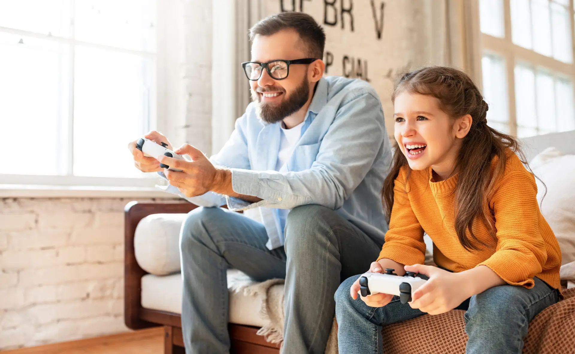 A man and a girl, both smiling and focused, are sitting on a couch playing video games with controllers—a perfect snapshot of what to do when your child is obsessed with video games. Sunlight streams through large windows, casting a warm glow on the girl’s yellow sweater as the man adjusts his glasses.