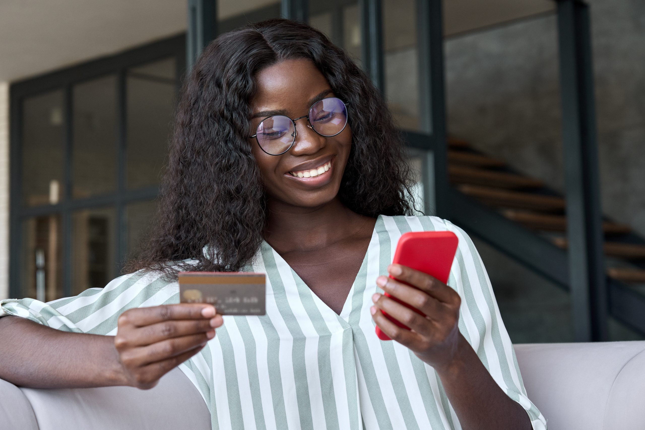 A woman with long curly hair and glasses sits on a couch, smiling at her phone. Dressed in a striped blouse, she holds a credit card, possibly shopping online. Stairs and glass panels are in the background, reminding us to be cautious of gift card scams while enjoying the convenience of digital shopping.