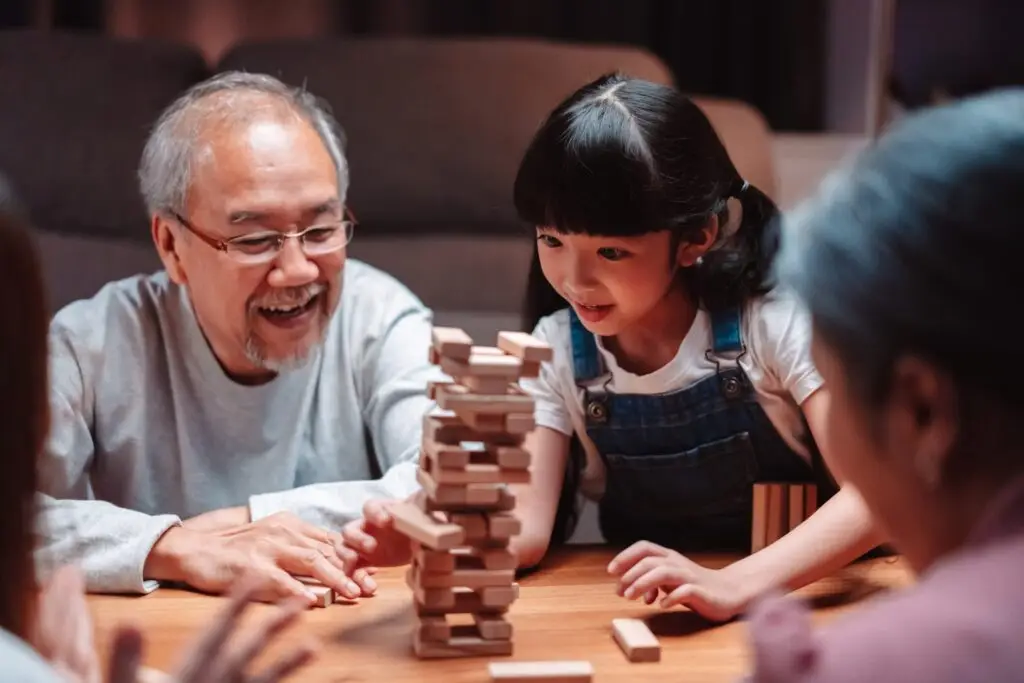 A grandfather and his granddaughter enjoy unplugging from technology by playing a game of Jenga with the family. The young girl, wearing a white shirt and denim overalls, carefully pulls a block from the tower as the grandfather watches with a smile.