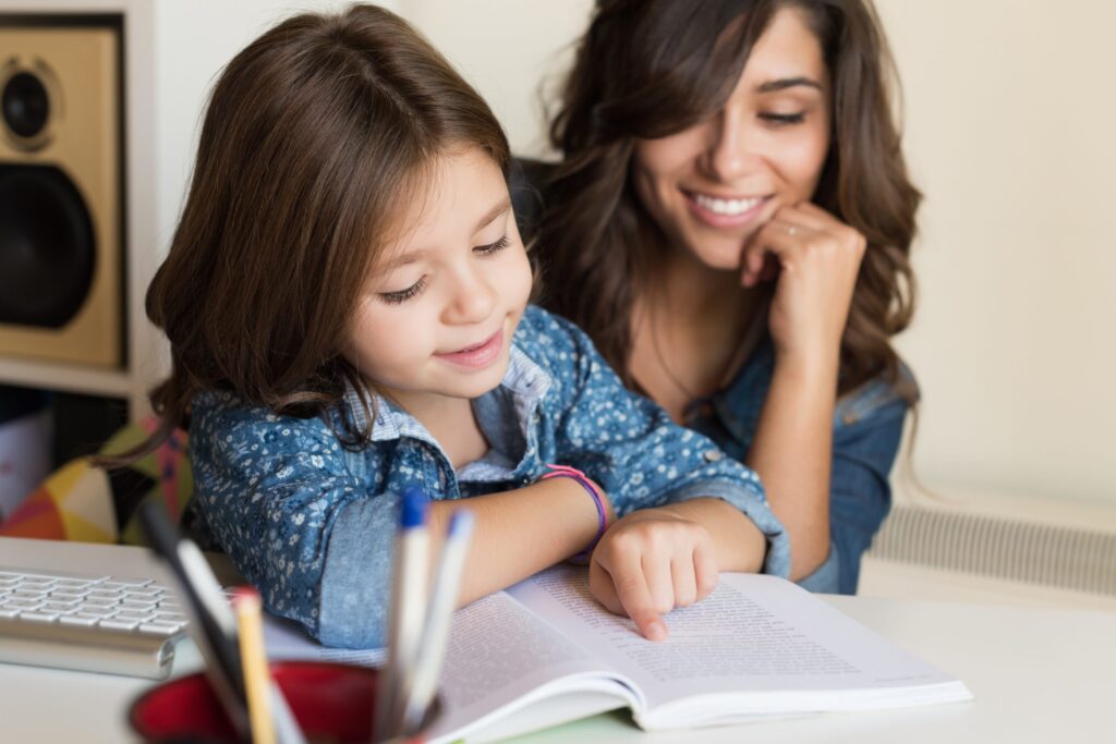 A young girl with long brown hair reads "Easy as A-B-C: How to Help Your Child with Reading," pointing at the text and smiling in her denim shirt. Beside her, a woman with wavy brown hair leans on her hand, observing warmly. They sit at a desk adorned with a keyboard and pencils.