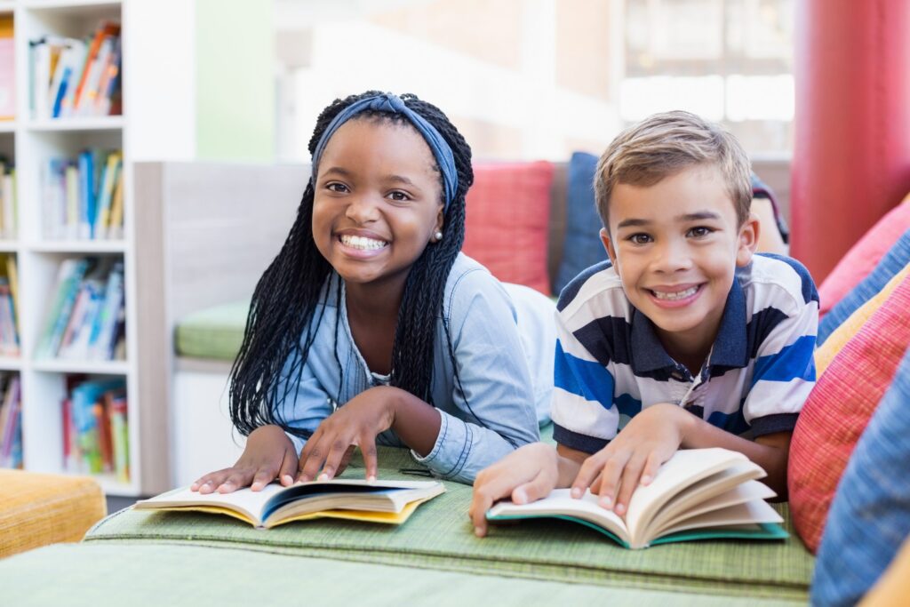 Two children lie on their stomachs, smiling and reading books in a cozy library setting, perfect for indoor educational activities. The girl, with braided hair, wears a light blue shirt, while the boy sports short hair and a striped polo. Shelves filled with books are in the background.