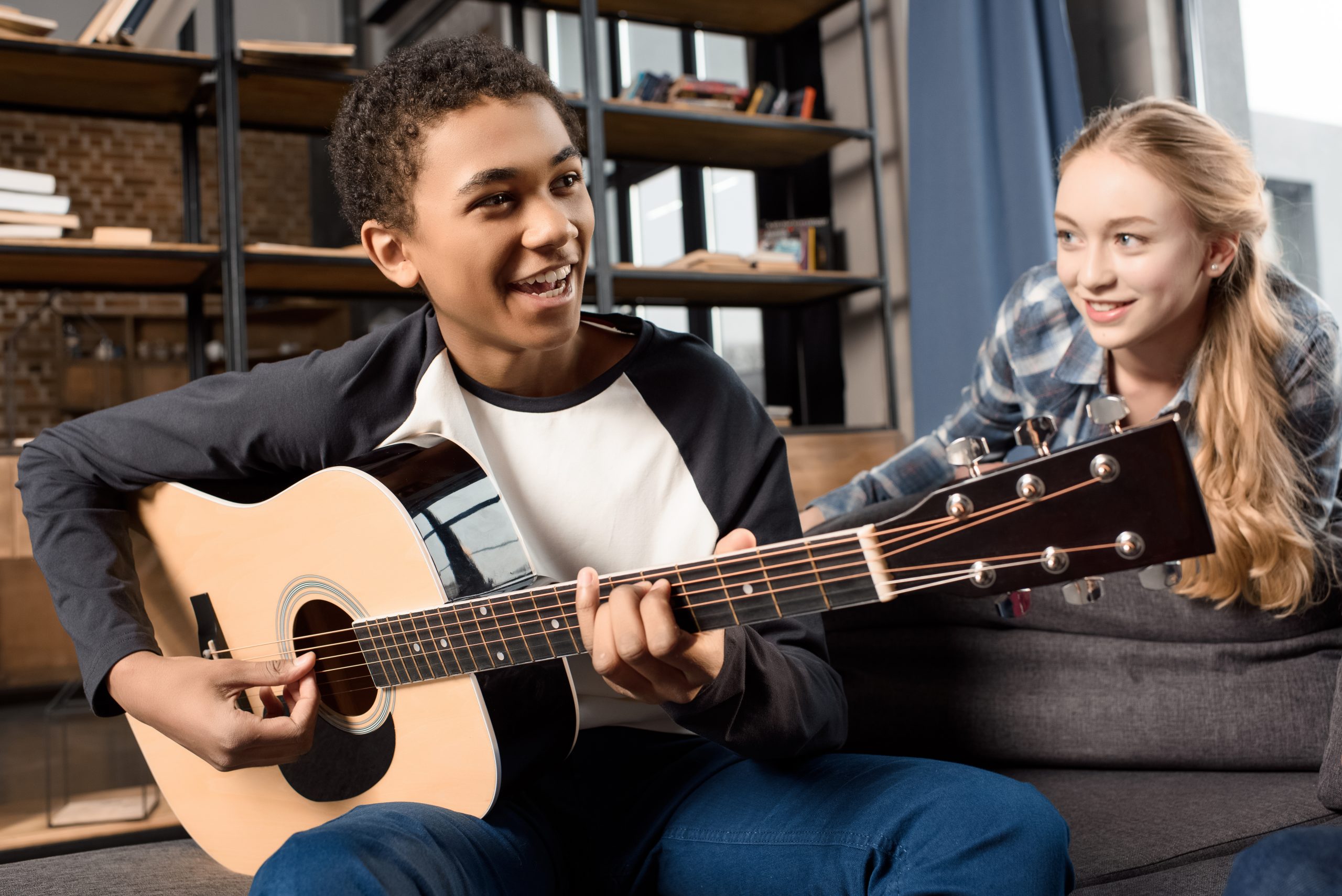 A boy is sitting on a couch, smiling while playing an acoustic guitar—a perfect escape as they embrace the idea of "enough is enough" by setting aside their phones. A girl next to him leans back, watching him with a smile. The room has shelves with books and a window in the background.