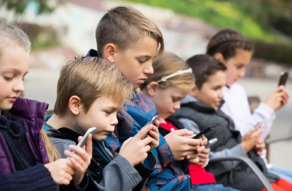 A row of young children sits on a bench outdoors, each deeply engaged with their smartphone. The scene, set against a blurred backdrop of greenery, subtly hints at why limiting screen time is important for kids.