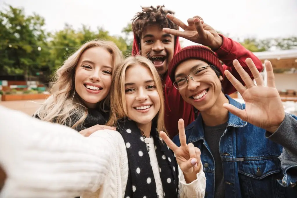 A group of four friends smiling and posing for a selfie outdoors—two in the front making peace signs, a third in the back mimicking their pose, while the fourth waves. With trees and a cloudy sky behind them, they're having fun as any user might ask themselves, "Is BeReal safe for kids?