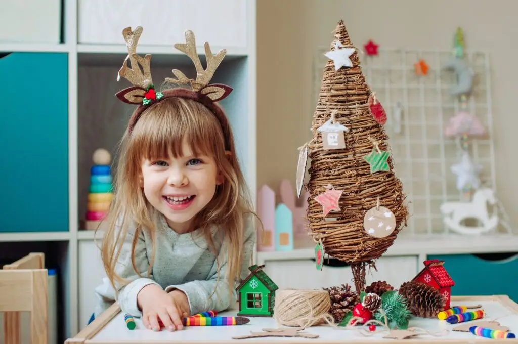 A smiling child wearing reindeer antlers leans on a table, surrounded by supplies from the "Five Easy Christmas Crafts for Kids." A homemade tree made of woven wood is adorned with small ornaments, while bright toys and colorful decorations fill the background.