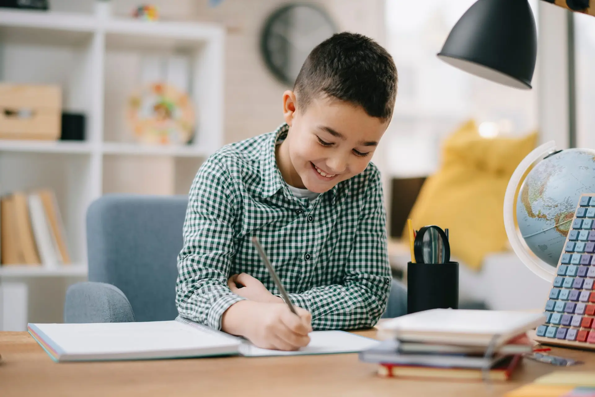 A young boy, inspired by how to motivate a child to do homework, sits at a desk in a cozy room, smiling as he writes in a notebook. The cluttered desk holds books, a globe, an abacus, and stationery. A lamp casts its glow over the space. Shelves and a yellow bean bag complete the inviting scene.