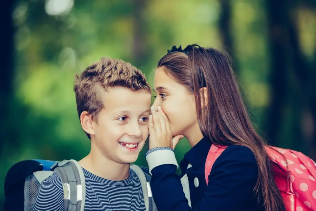 A girl whispers into a smiling boy's ear. Both children wear backpacks and stand outdoors, surrounded by blurred greenery, suggesting a park or forest setting—where they might enjoy one of the 10 games to play with friends that don’t require anything.