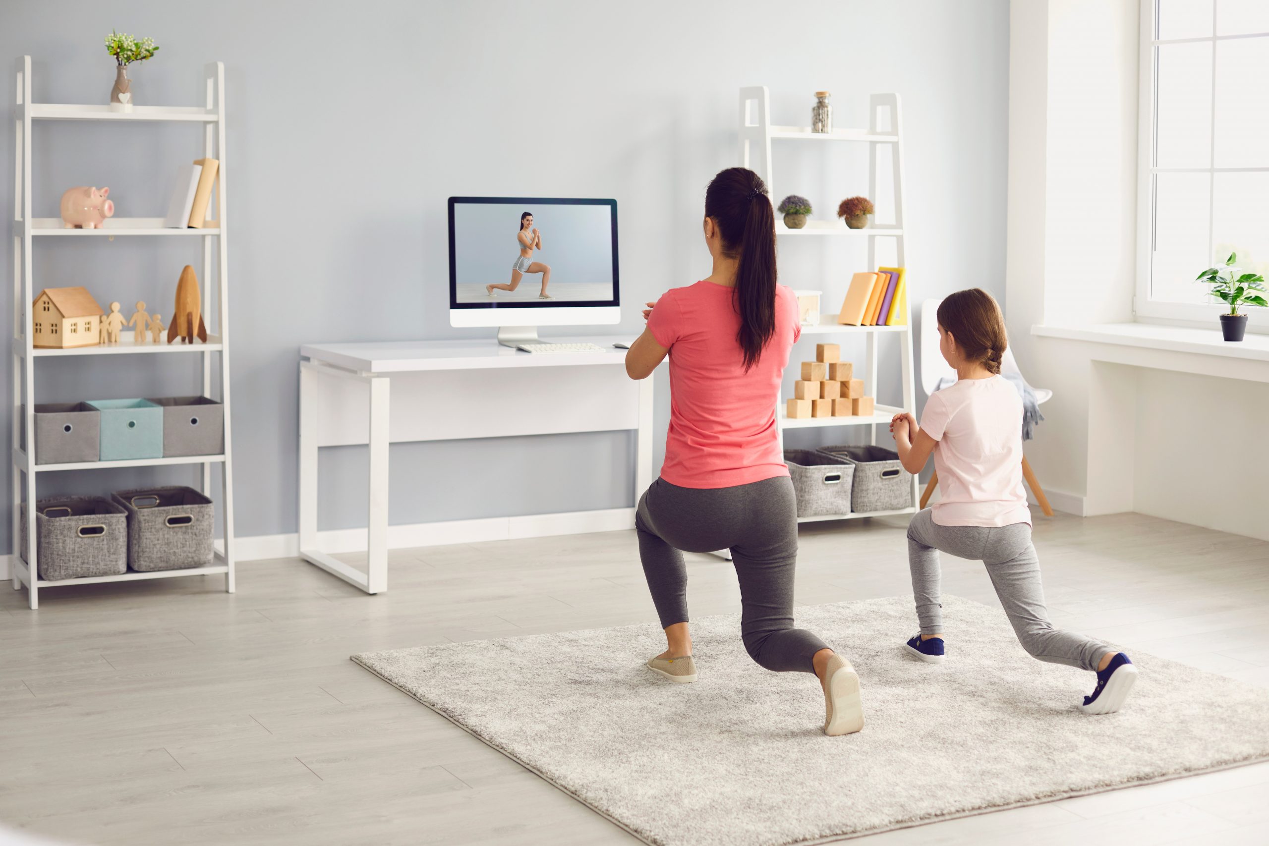 A woman and child are exercising at home, following a workout video on a computer screen titled "Break a Sweat: Best TV Workout Apps." They perform lunges on a light gray carpet in the bright room, with shelves, bins, and a plant enhancing the inviting atmosphere.