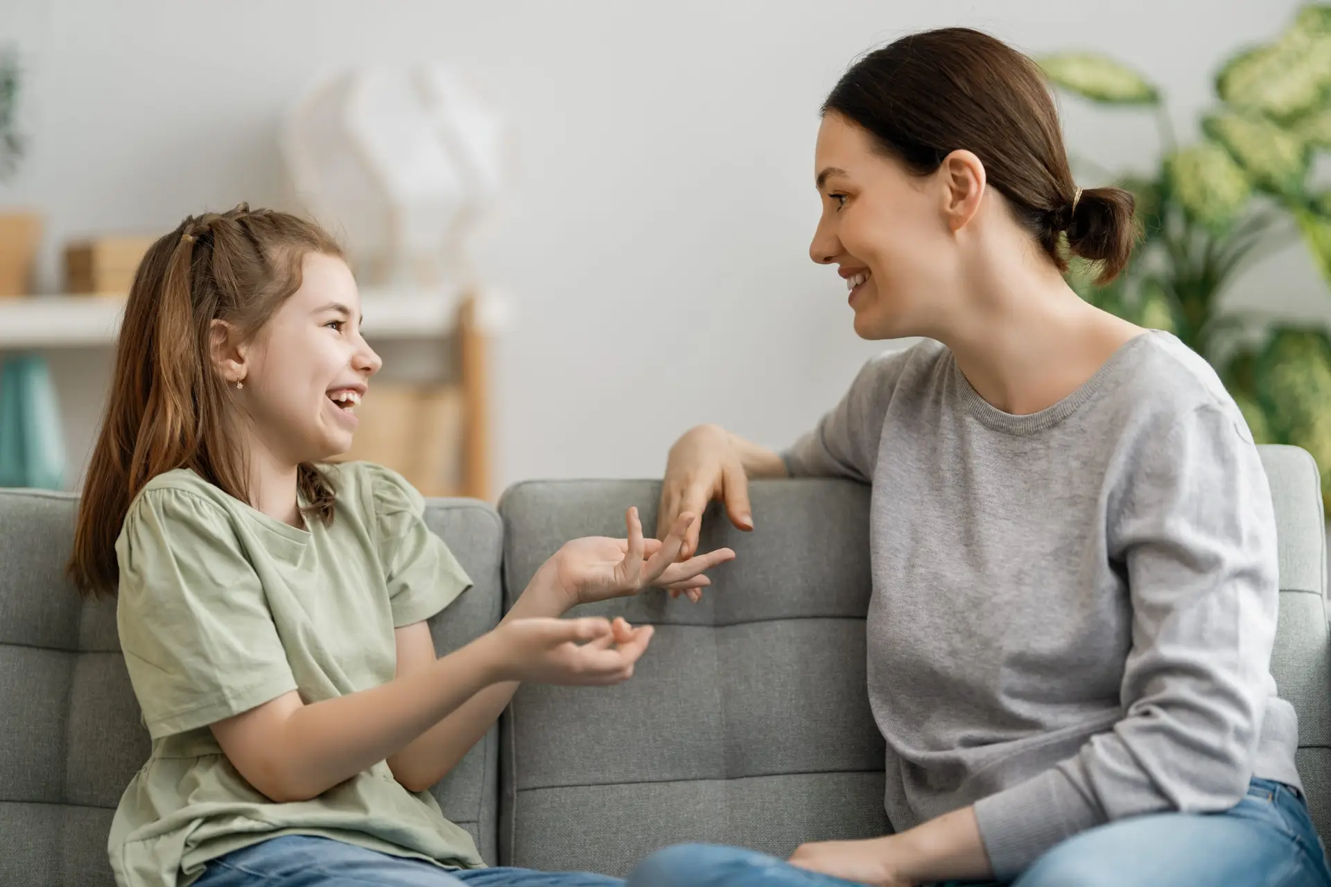 A woman and a young girl are sitting on a gray sofa, sharing smiles and lively chat. The woman, with dark hair tied back, gently guides the conversation to nurture the girl's social-emotional skills in their cozy room.