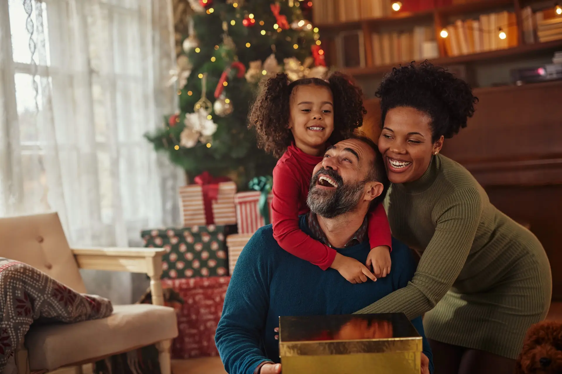 A joyful family, including a man, woman, and child, celebrates the holidays in a cozy living room. They are smiling and holding a golden gift in front of a beautifully decorated Christmas tree with wrapped presents underneath, embracing one of their eighteen cherished Christmas traditions.