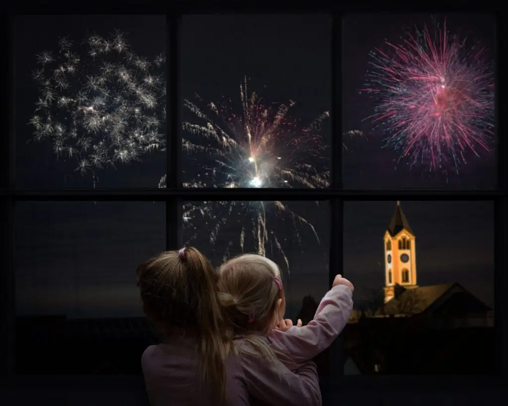 Two children, seen from behind, enjoy colorful fireworks through a window at night. A clock tower is illuminated in the background by vibrant explosions. One child points toward the display, capturing the perfect moment for New Year’s Eve at home with family.