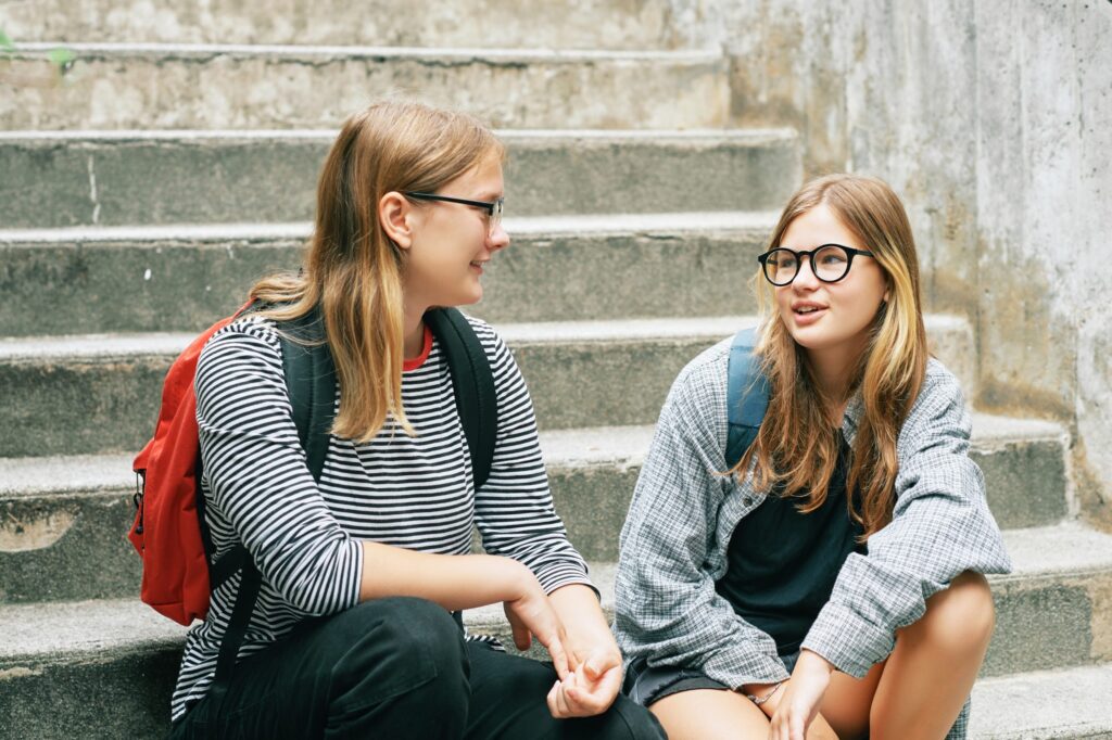 Two young women sit on concrete steps, laughing and chatting about "Ten Things Every 13-Year-Old Should Know." Both wear glasses and backpacks. One sports a striped shirt, the other a plaid one. The casual, urban setting adds charm to their lively discussion.