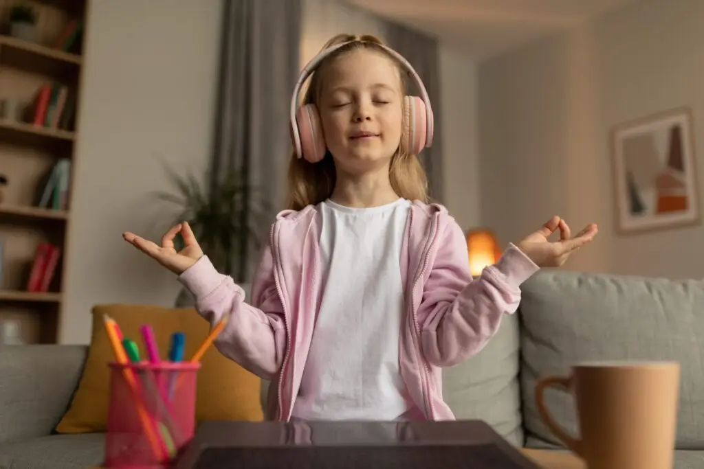 A young girl in a pink hoodie and white shirt sits on a couch with her eyes closed, hands in a meditative pose. Her pink headphones hint at the surprising benefits of being bored. Nearby, a laptop, colorful pens, and a mug sit ready for when inspiration strikes.