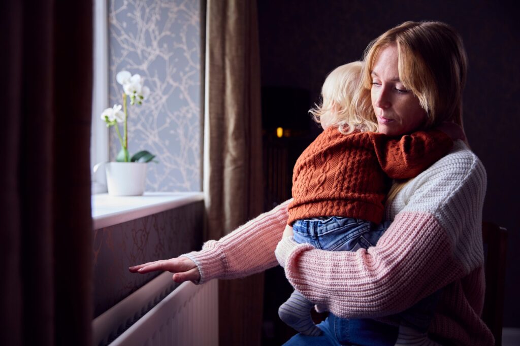 A woman in a cozy sweater holds a young child, cuddled in an orange sweater and jeans, as she gently touches the radiator for warmth. A potted plant adorns the softly lit room, offering calm—a serene moment amidst winter blues often mistaken for seasonal affective disorder.