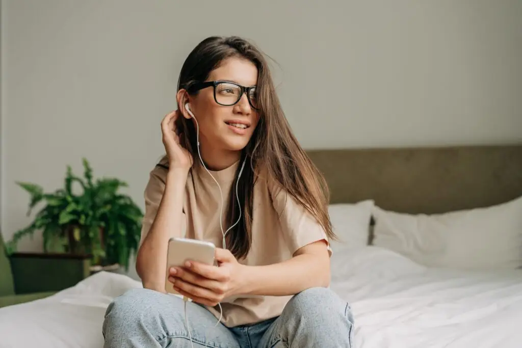 A young woman with long brown hair and glasses sits on a bed, holding her smartphone while pondering TikTok safety concerns. Dressed in a beige shirt and jeans, she listens through earphones. A green plant is visible in the background as she smiles and looks away from the camera.