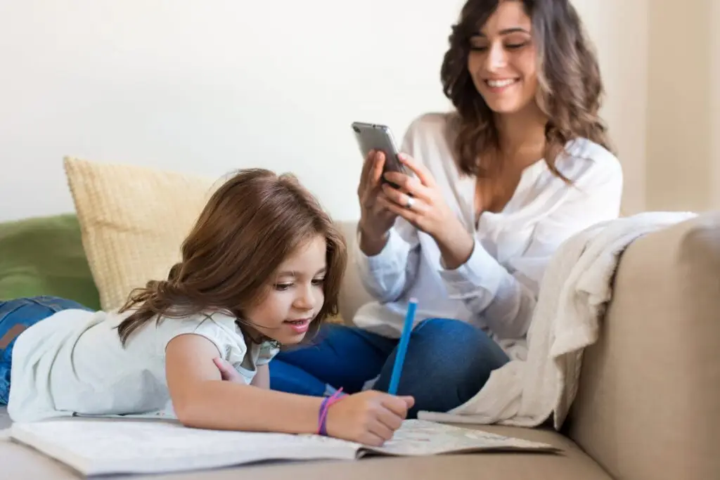 A young girl lies on the couch drawing in a notebook with a pencil. Next to her, a woman smiles at her phone, possibly exploring what parental control apps are and how they work. Both appear relaxed in a cozy living room with natural light pouring in.