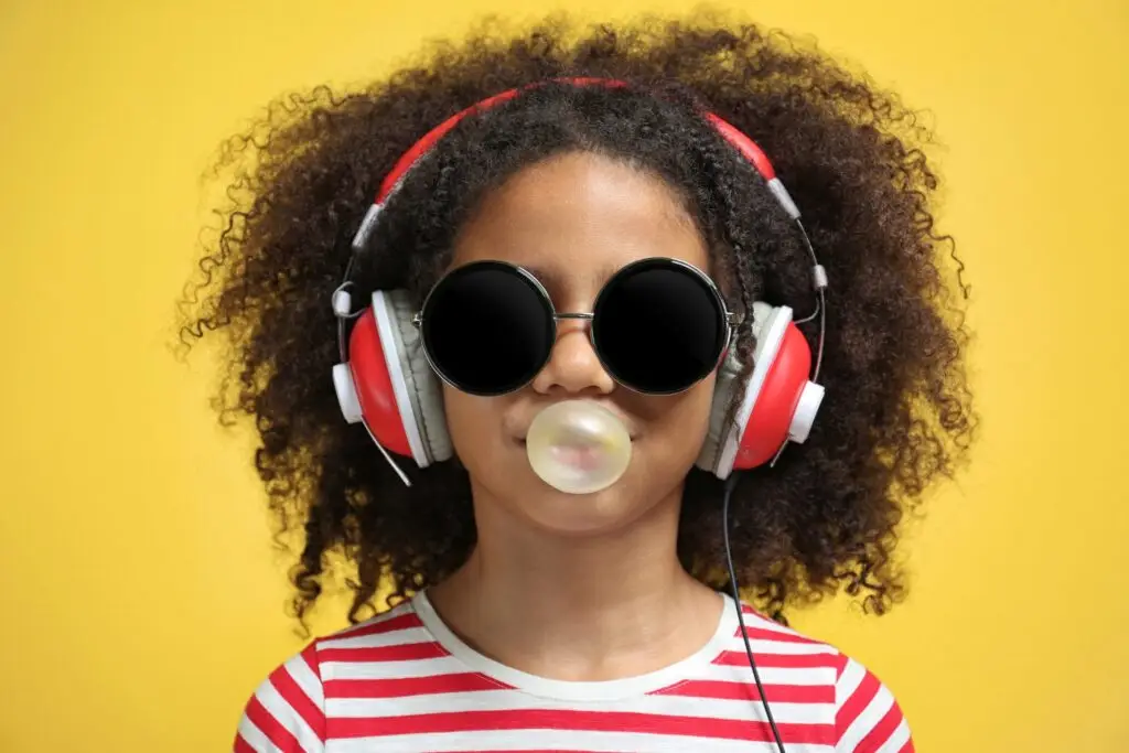 A young girl with curly hair wears round black sunglasses and red headphones, blowing a bubble with gum. She showcases the playful charm of "Gum for Kids: The Dos and Don’ts" in her stylish red and white striped shirt against a bright yellow background.