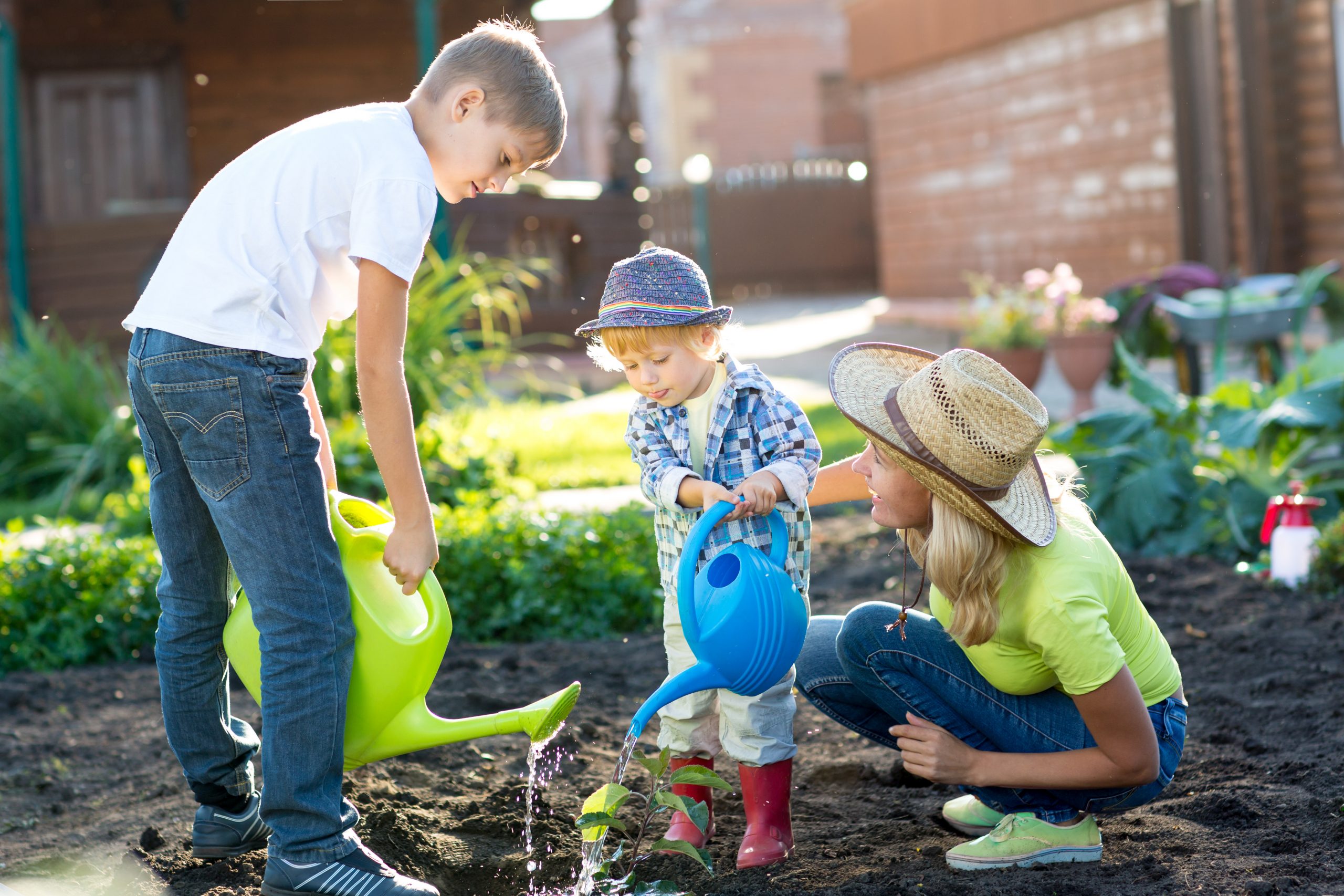 A woman and two children are gardening, embracing a chance for independence. The woman and one child, both wearing hats, bend down to water plants with colorful watering cans. The ground is freshly tilled and dark, with a wooden building in the background.