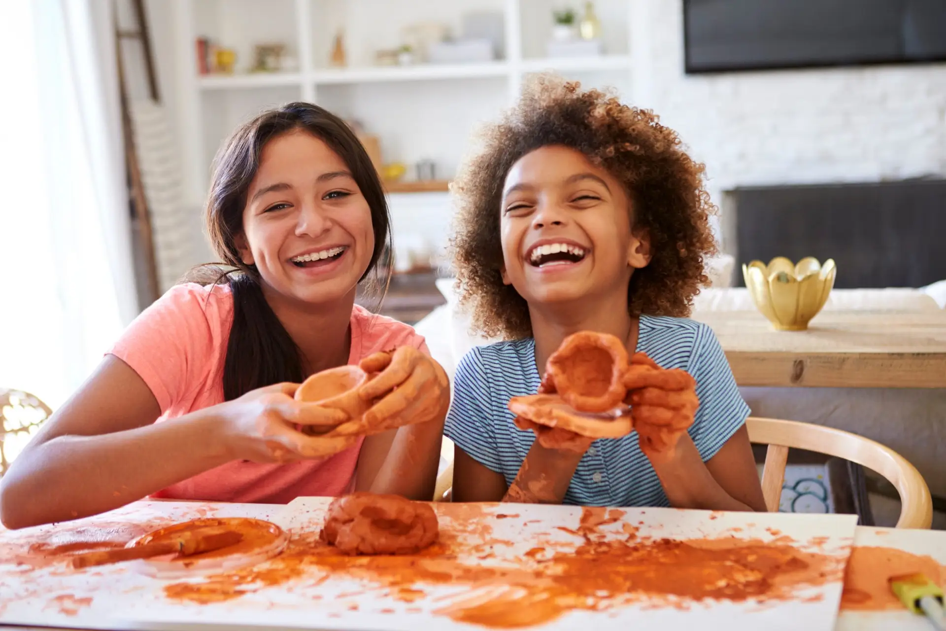 Two children smiling while playing with clay at a table, discovering how to help your child make friends through creativity. The girl on the left wears a pink shirt, and the one on the right dons blue stripes. Their hands dance in orange clay, with a cozy living room in the background.