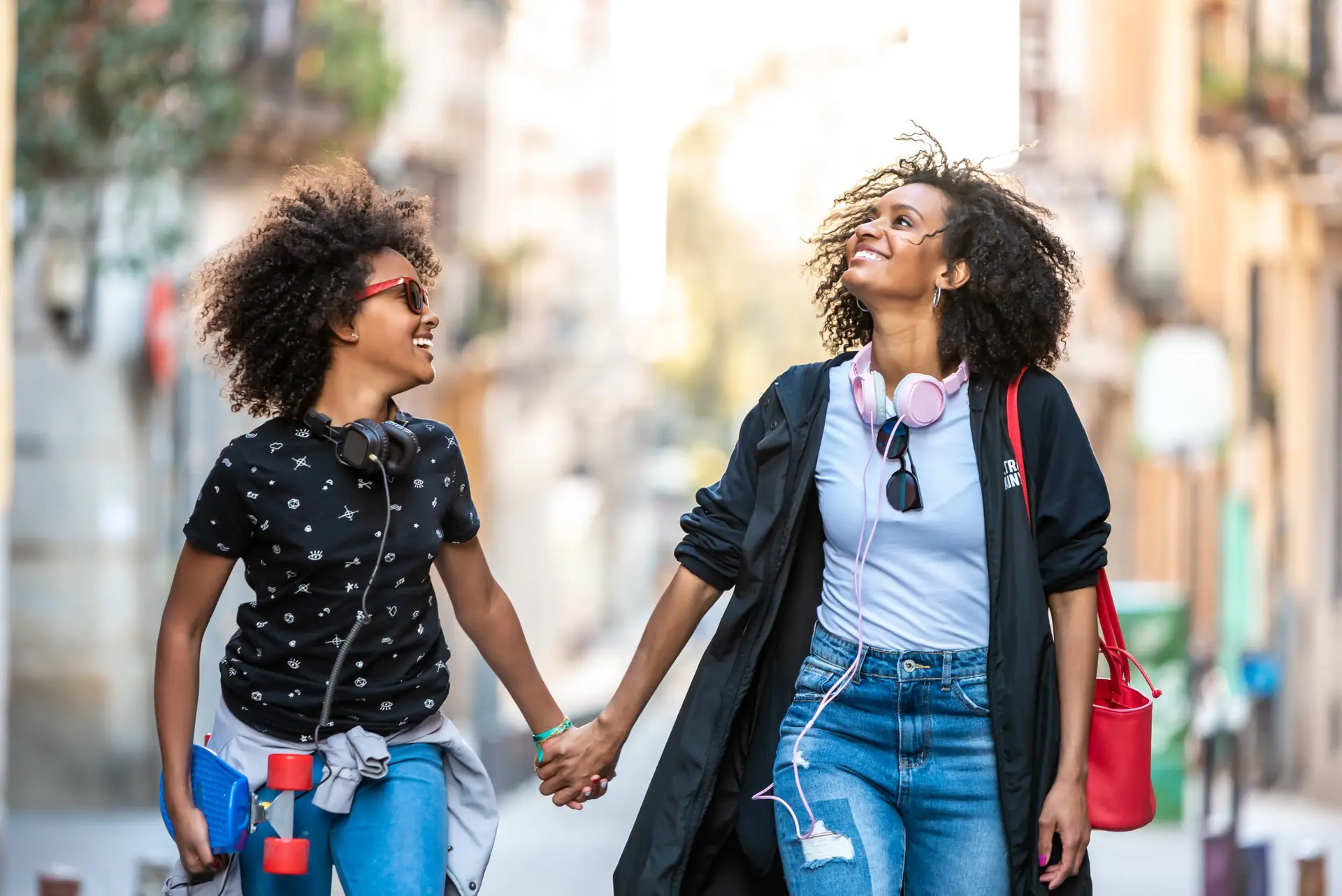 Two people with curly hair walk hand in hand down a sunlit street, engaging in one of the fifty fun activities to do with your teenager. They smile at each other as one wears red sunglasses and headphones, while the other has pink headphones around their neck and carries a red bag. A skateboard hangs from one person's backpack.