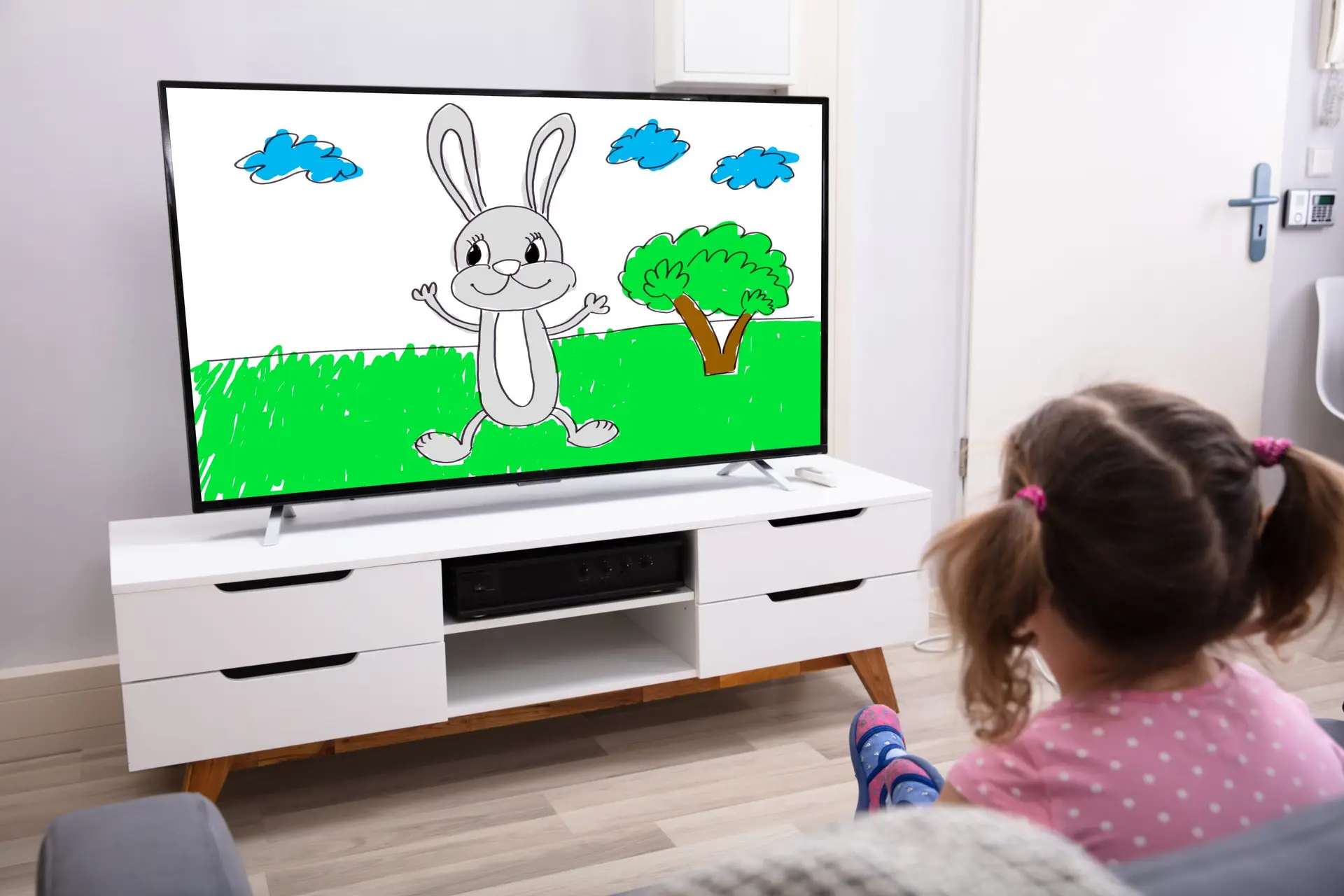 A young girl with pigtails sits on the floor, exploring the pros and cons of kids watching TV as she enjoys a cartoon featuring a smiling rabbit on a modern flat-screen. The room is bright and minimally furnished, with a white TV stand and wooden flooring.