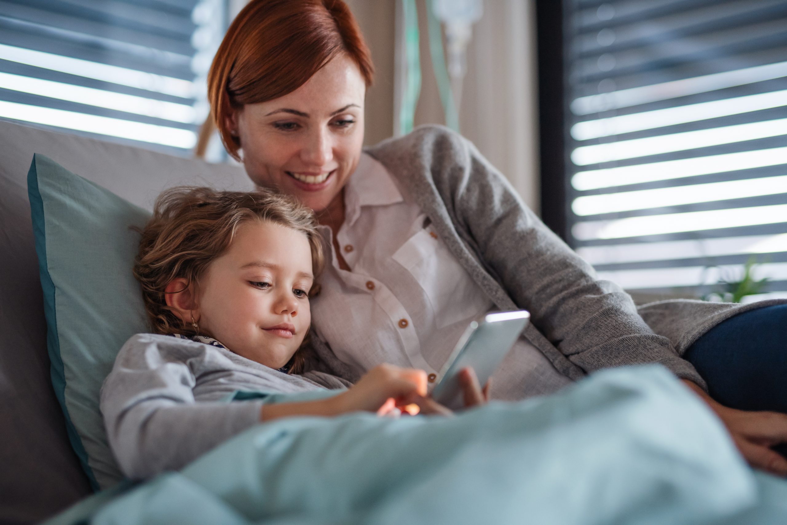 A smiling woman and a young child lie on a bed, looking at a smartphone together. With red hair and curly hair respectively, they exude relaxation and joy in the sunlit room with blinds. It's heartwarming to see them bond over being safe sleuths while exploring the child's phone together.