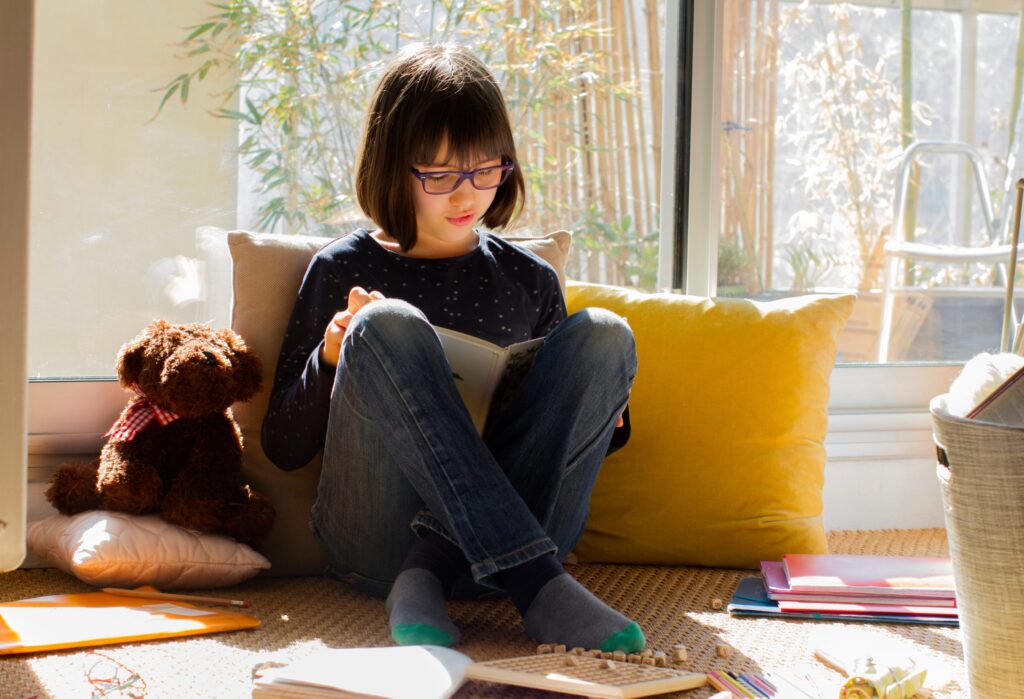 A young girl with glasses sits by a sunlit window, reading a book on "Leaving Your Child Home Alone: What You Should Know." Surrounded by pillows, a plush teddy bear, and papers scattered on the floor, the scene is cozy and bright, with bamboo visible outside.