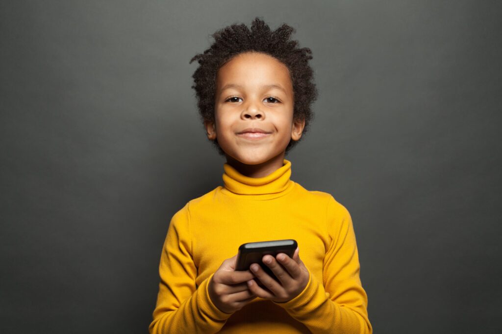 A child with curly hair wearing a yellow turtleneck holds a smartphone and smiles, as if they've mastered the art of following the top ten cell phone rules for kids, standing confidently against a dark gray background.