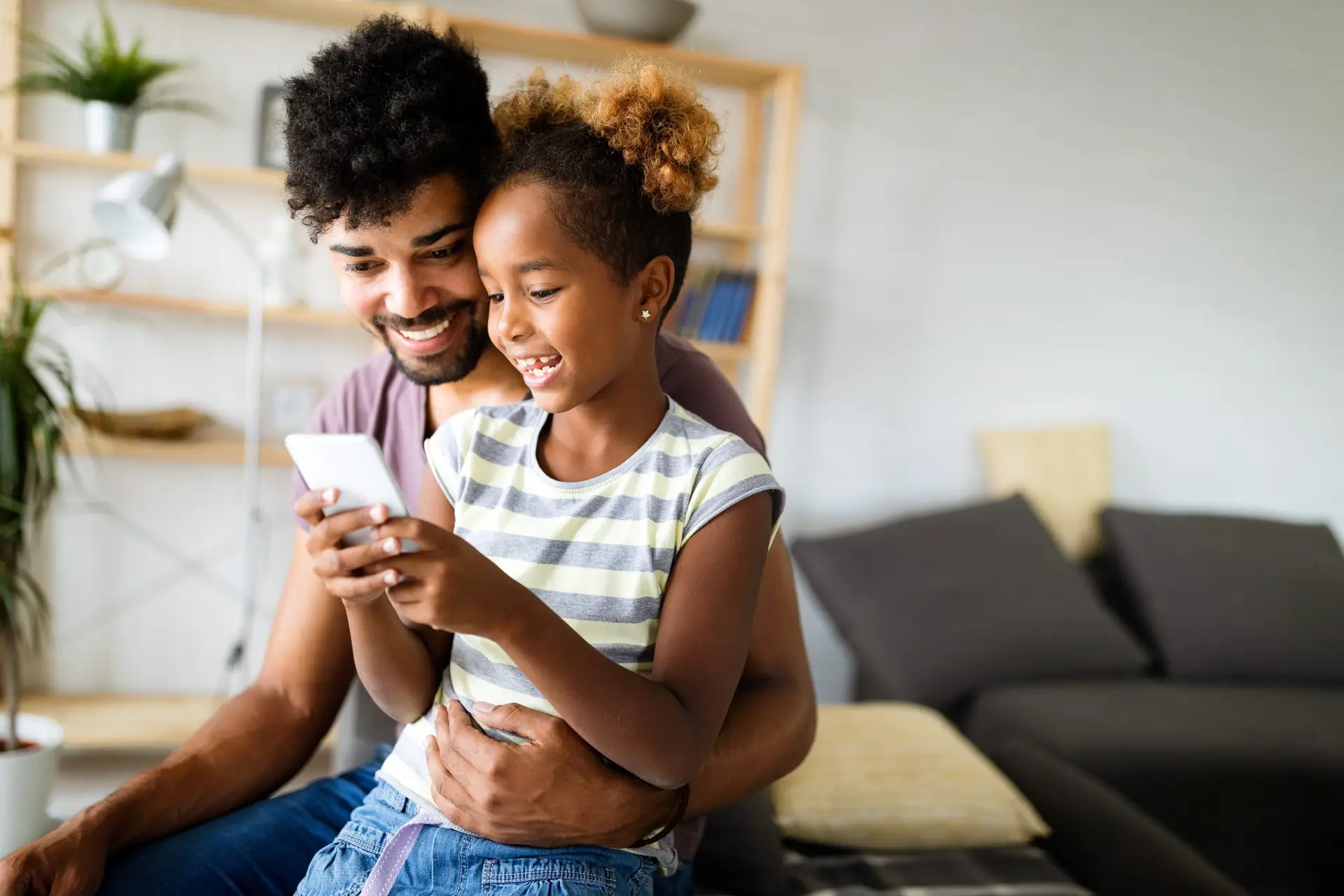 A man and a young girl are sitting together on a couch, smiling while looking at a smartphone. The room has shelves and plants in the background, creating a cozy atmosphere, prompting the question: when should I give my child a phone?