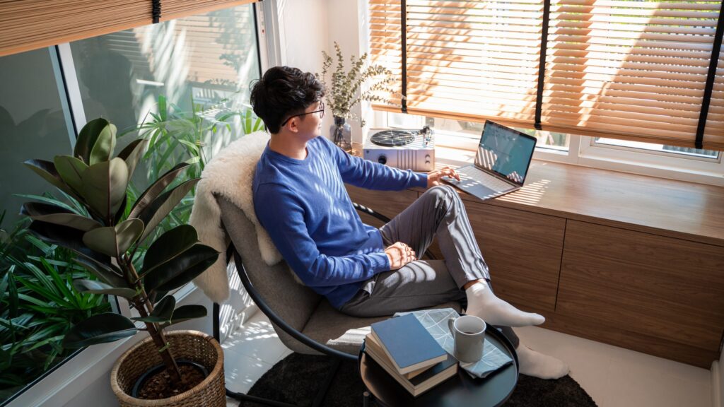 A person in a blue sweater and glasses sits in a cozy room, using a laptop on a windowsill—the perfect scene from "Fifteen Home Office Tech Essentials." Sunlight streams through blinds, illuminating a large plant and books on the table. A cup and small speaker accentuate the ambiance.