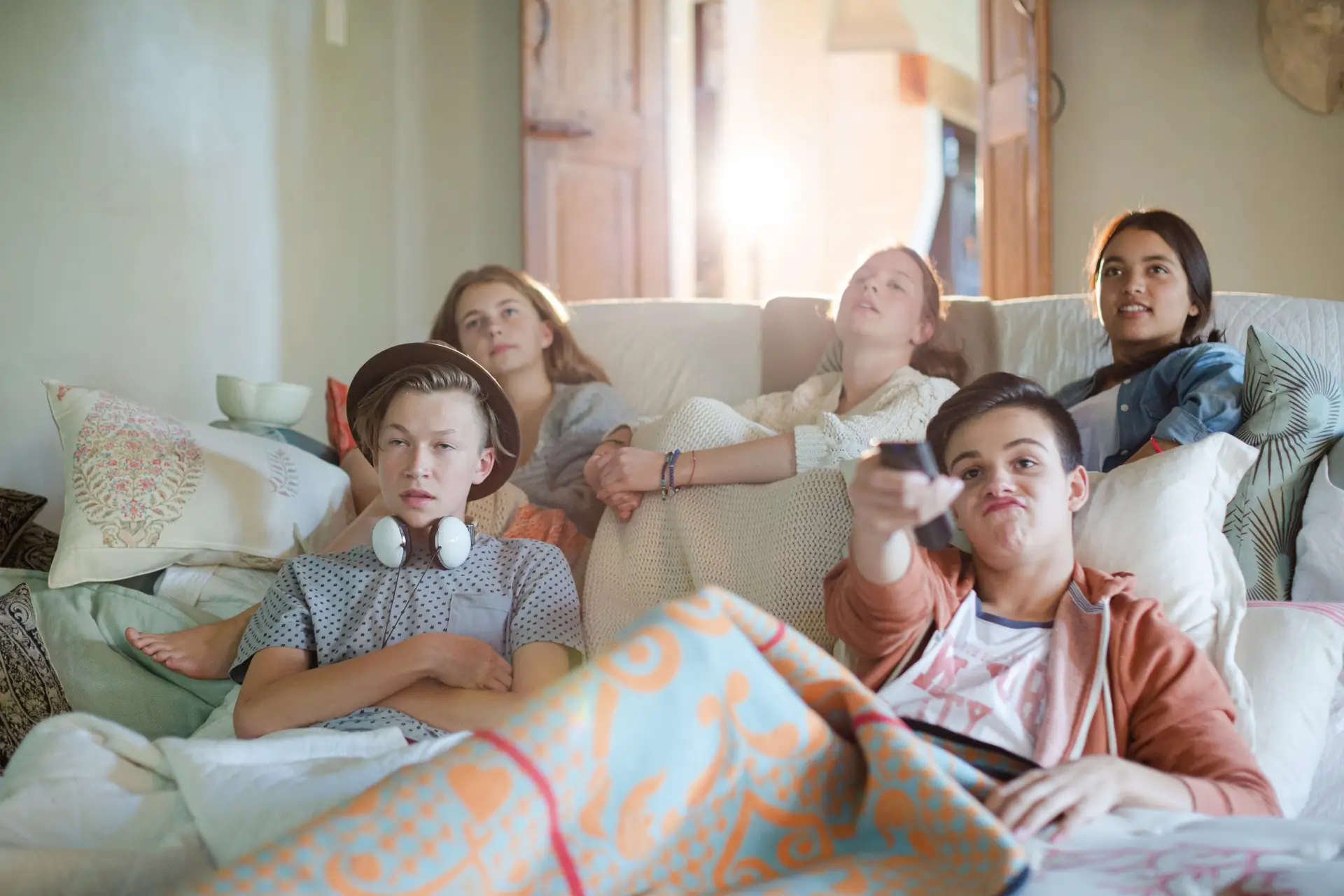 Five teenagers sit on a couch, wrapped in blankets and engrossed in one of the "Twelve Good TV Shows for Tweens & Teens." One holds a remote control while another has headphones around their neck. They appear relaxed and intently focused on the screen.