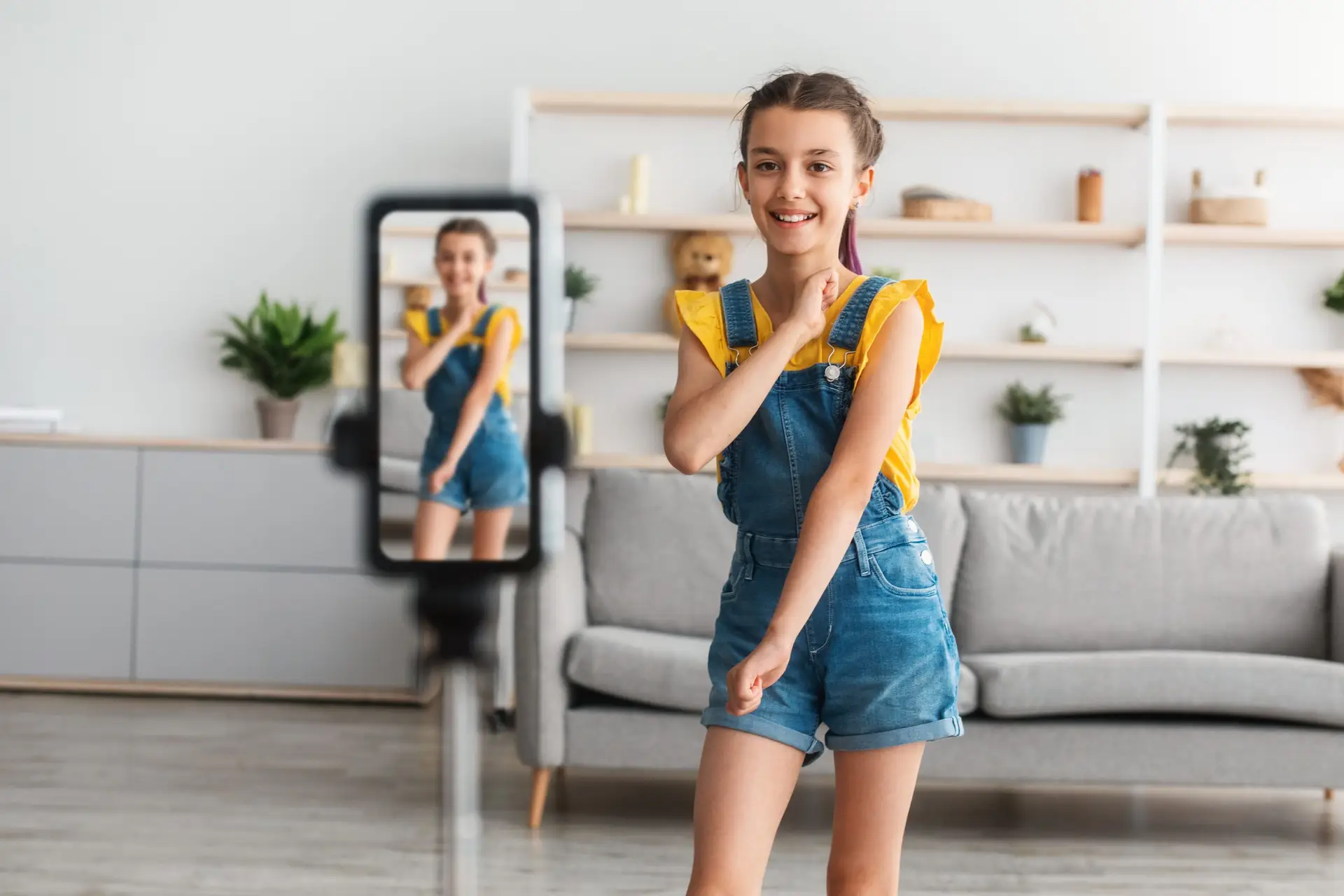 A young girl in a yellow top and denim overalls beams at a phone on a tripod, likely creating content for TikTok for Kids. She stands in a modern living room with minimalist decor, featuring a gray sofa and plants neatly arranged on shelves.