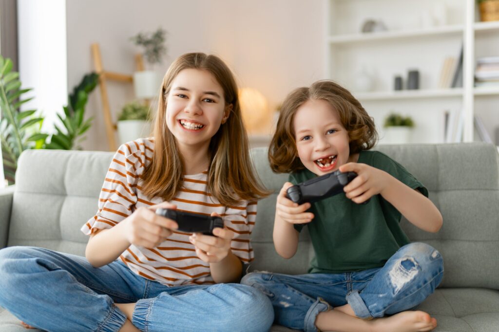 Two children sit on a couch, laughing and holding video game controllers. They seem to be having a blast playing together in their cozy room adorned with plants and shelves. Considering games like Undertale for kids? It's worth exploring what's popular among young gamers to ensure it's appropriate.