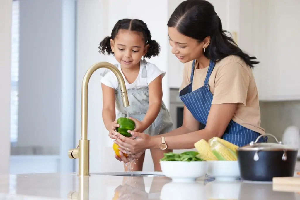 A woman and a young girl stand at a kitchen sink, washing vegetables together as she subtly teaches responsibility. The girl holds a green pepper under the running water while the woman assists. Nearby, corn cobs and leafy greens are scattered on the counter, waiting to be cleaned.