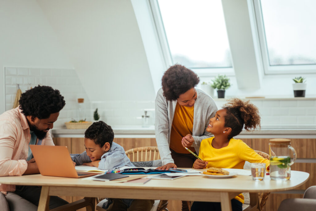 A family of four sits at a kitchen table. Two adults help two children with art and schoolwork, showing How to Motivate a Child to Do Homework. Papers, notebooks, colored pencils, snacks, and drinks are spread across the table.